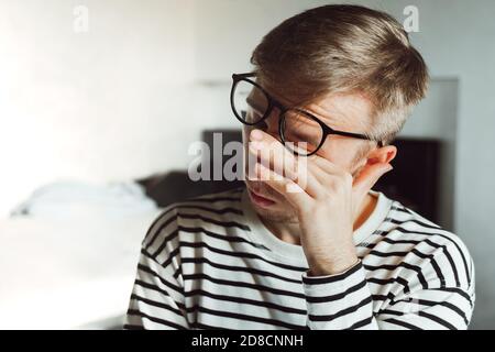 Young man in eyeglasses feeling exhausted, looking tired after reading, Frustrated millennial rubbing eyes in home office. Sight loss, blurry vision, Stock Photo