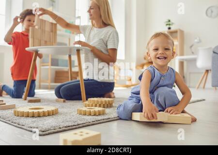 Cute little kid looking at camera and smiling while elder brother and mum are playing in background Stock Photo