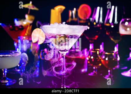 Close up of dry Martini drink with olives at bar counter on a cocktail bar background. Colorful various drinks in a bar with dry ice smoke effect.