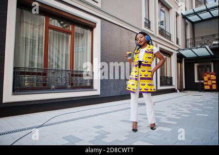 Stylish african american women in yellow jacket posed and earphones on street against modern building with hot drink in disposable paper cup. Stock Photo