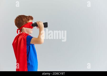 Little boy in a mask and cloak of a superhero looks through binoculars on a gray background. Stock Photo