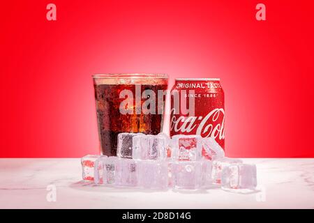 London, United Kingdom - October 29 2020:  Ice cold can of Coke sits next to glass of soda with ice cubes on a red background Stock Photo