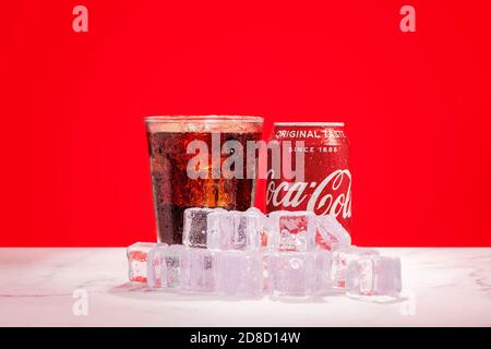 London, United Kingdom - October 29 2020:  Ice cold can of Coke sits next to glass of soda with ice cubes on a red background Stock Photo