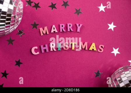 Merry Christmas greeting words made of wooden letters, on a purple background decorated with stars confetti, and mini disco balls. Stock Photo
