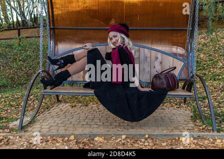 A beautiful woman sits on a garden swing, forged in a burgundy coat and biret, an adult smiles at the camera, in the fall against the backdrop of a Stock Photo