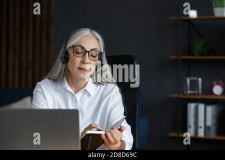 Portrait of beautiful smiling elderly woman studying online. Online education, remote working, home education. Grey-haired senior woman writing in Stock Photo
