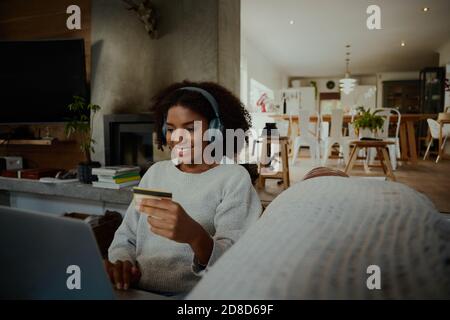 African young woman with credit card in hand sitting with laptop and making transfer money and listening to music Stock Photo