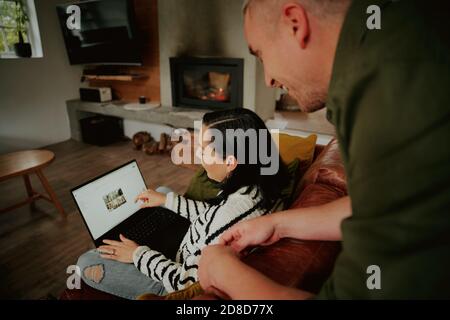 Young woman sitting on couch showing screen to husband while browsing on laptop Stock Photo