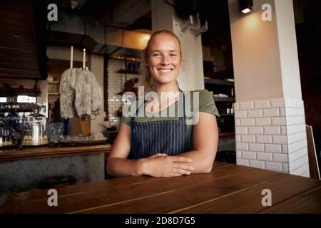 Portrait of smiling young female waitress wearing apron standing leaning on table in cafe Stock Photo