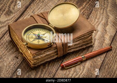 retro leather-bound journal with decked edge handmade paper pages, antique brass compass and a stylish pen on a rustic wooden table, journaling concep Stock Photo