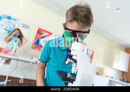 Boy wearing face mask and protective glasses using microscope in laboratory Stock Photo