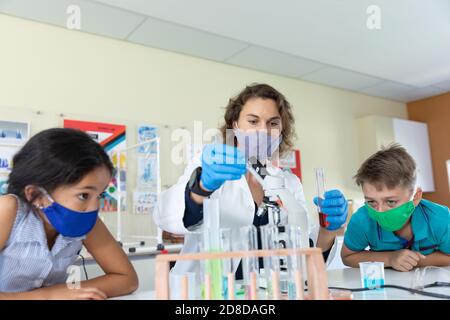 Female teacher wearing face mask and protective glasses teaching students in laboratory Stock Photo