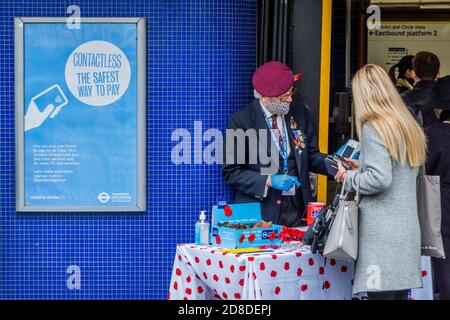 London, UK. 29th Oct, 2020. It is London Poppy Day and a veteran (in the high risk covid age group) of Airborne forces, in his red beret, collects for the Royal British Legion, as a steady flow of passengers exits Sloane Square Tube Station. He wears a mask and gloves and mostly collects funds using contactless payments (although he still has a 'tin' for cash). Travellers mostly wear masks after they become mandatory. Credit: Guy Bell/Alamy Live News Stock Photo