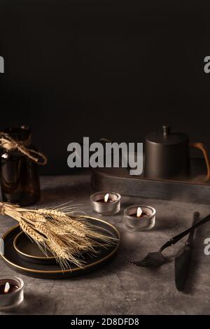 Vintage template with old metal tableware, ear of wheat and lit candles on black stone background for concept design. Fall concept. Interior design concept. Stock Photo