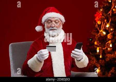 Bearded Santa Claus in eyeglasses holding credit card and paying online using his mobile phone Stock Photo