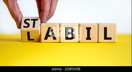 Hand turns a cube and changes the German word 'labil' - unstable in English to 'stabil' - stable in English. Beautiful yellow table, white background, Stock Photo