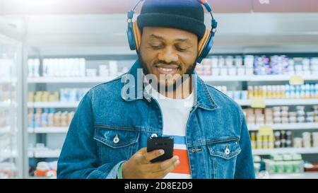 At the Supermarket: Portrait of a Stylish African American Guy with Headphones, Using Smartphone, Walks Through Frozen Goods Section of the Store. Stock Photo