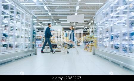 At the Supermarket: Man in a Hurry Pushes Shopping Cart full of Items, He's Walking Through Different Section of the Big Bright Mall. Stock Photo