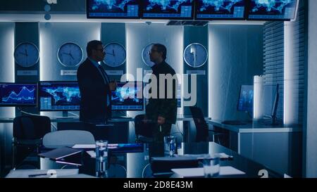 Federal Special Agent Talks To Military Man in the Monitoring Room. In the Background Busy System Control Center with Monitors Showing Data Flow. Stock Photo