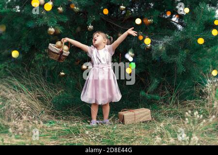 cute little girl in a pink dress decorates a Christmas tree. the child found a gift under the tree and emotionally raised her hands up. new year and c Stock Photo