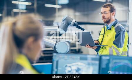 At the Factory: Female Mechanical Engineer Designs 3D Engine on Her Personal Computer, Male Automation Engineer Uses Laptop for Programming Robotic Stock Photo