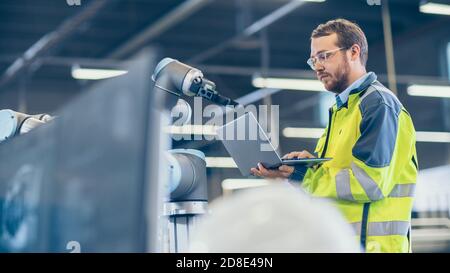 At the Factory: Automation Engineer Uses Laptop for Programming Robotic Arm. New Era in Automatic Manufacturing Industry. Stock Photo