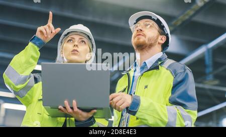 Male and Female Industrial Engineers Work on a Manufacturing Plant, They Discuss Project, Point in the Direction of the Machinery while Using Laptop Stock Photo