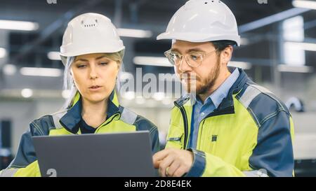Male and Female Industrial Engineers Work on a Manufacturing Plant, They Discuss Project, Point in the Direction of the Machinery while Using Laptop. Stock Photo