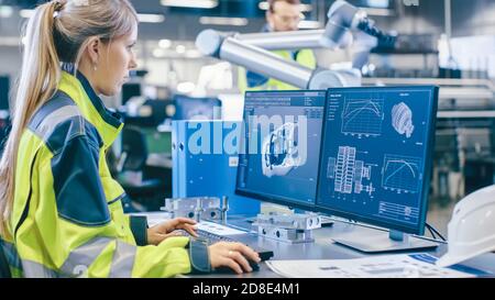 At the Factory: Female Mechanical Engineer Designs 3D Engine on Her Personal Computer, Male Automation Engineer Uses Laptop for Programming Robotic Stock Photo
