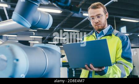 At the Factory: Automation Engineer Uses Laptop for Programming Robotic Arm. New Era in Automatic Manufacturing Industry. Stock Photo