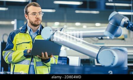 At the Factory: Automation Engineer Uses Laptop for Programming Robotic Arm. New Era in Automatic Manufacturing Industry. Stock Photo