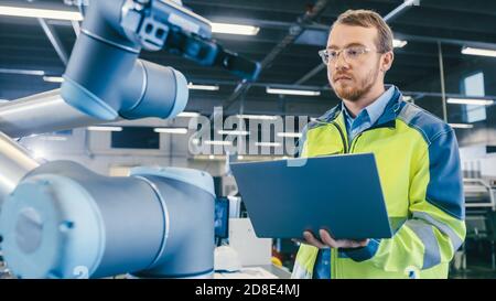 Low Angle Shot At the Factory: Automation Engineer Uses Laptop for Programming and Testing Robotic Arm. New Era in Automatic Manufacturing Industry. Stock Photo