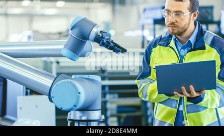 At the Factory: Automation Engineer Uses Laptop for Programming Robotic Arm. New Era in Automatic Manufacturing Industry. Stock Photo