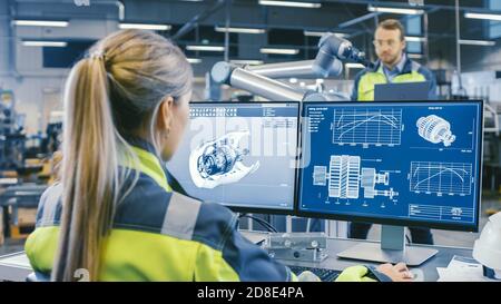 At the Factory: Female Mechanical Engineer Designs 3D Engine on Her Personal Computer. In the Background Male Automation Engineer who Uses Laptop for Stock Photo