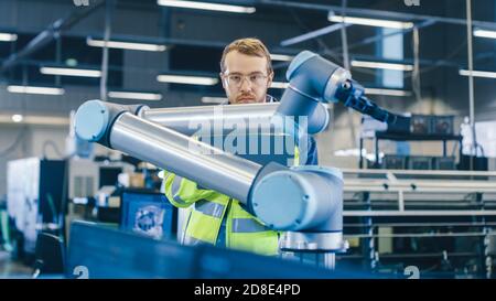 At the Factory: Automation Engineer Uses Laptop for Programming Robotic Arm. New Era in Automatic Manufacturing Industry. Stock Photo