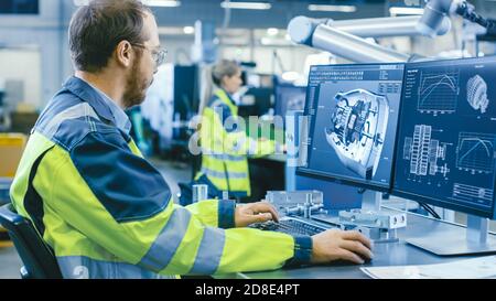 At the Factory: Mechanical Engineer Works on Computer, Designs in CAD 3D Model of the Engine. In the Background Factory Workers and Manufacturing Stock Photo