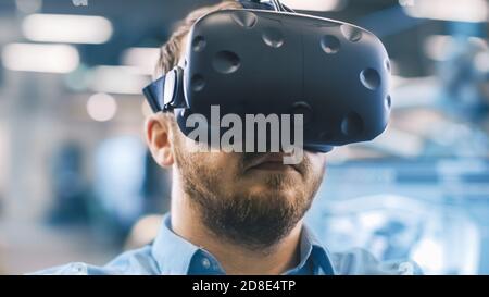 Portrait Shot of the Industrial Engineer Wearing Virtual Reality Headset and Gesturing with Controllers. In the Background Out of Focus Displays and Stock Photo
