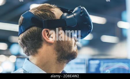 Portrait Shot of the Industrial Engineer Wearing Virtual Reality Headset and Gesturing with Controllers. In the Background Out of Focus Displays and Stock Photo