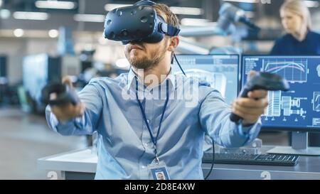 Portrait Shot of the Industrial Engineer Wearing Virtual Reality Headset and Using Controllers, ready to Work. In the Background Manufacturing Plant Stock Photo