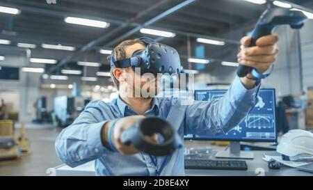 Portrait Shot of the Industrial Engineer Wearing Virtual Reality Headset and Using Controllers, ready to Work. In the Background Manufacturing Plant Stock Photo