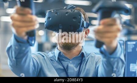 Portrait Shot of the Industrial Engineer Wearing Virtual Reality Headset and Using Controllers, ready to Work. In the Background Manufacturing Plant Stock Photo