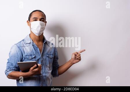 Latino man with facial mask holds a tablet and points to the empty space on white background. Social distance. Stock Photo