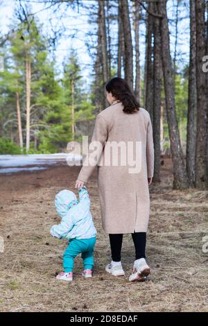 Mother with her little baby walking in the forest, toddler trying to step with legs on earth, mom holds hand Stock Photo