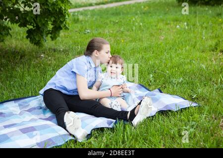 Teen age girl kissing her toddler sister while sitting on blanket in green grass on picnic, summer meadow, two girls Stock Photo