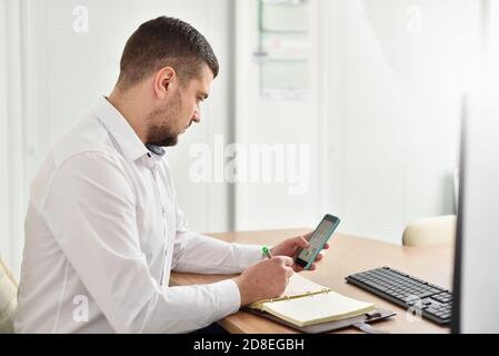 Young male office manager in the office looks carefully at the smartphone and writes in the notebook Stock Photo