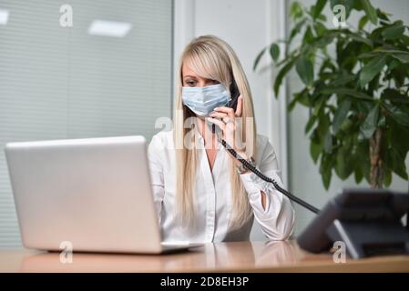 Young blonde business woman in a face mask talking on the phone in the office and working with a laptop Stock Photo