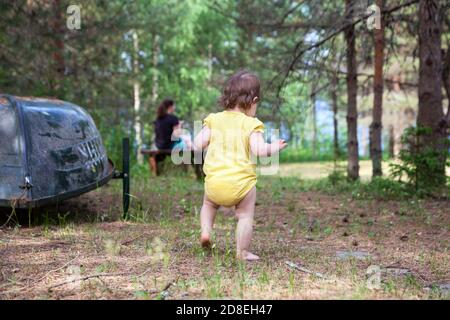 Baby girl running barefoot on earth in evergreen forest, Caucasian child comes to mother sitting on bench Stock Photo