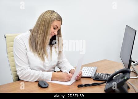 Blonde woman in the office talking on the phone and writing in a notebook Stock Photo