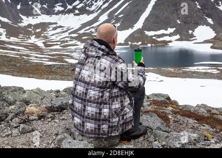 Backpacker resting with coffee thermos mug in mountains, adult man sitting on stones with picturesque view at mountain lake with glacier Stock Photo