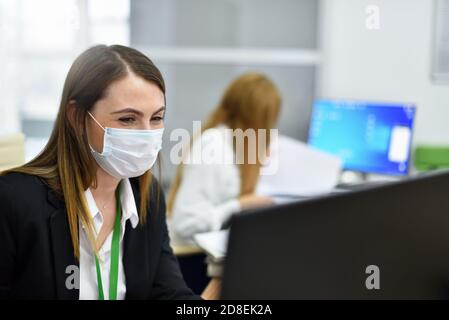 Happy woman office manager in protective mask works at computer in modern office Stock Photo